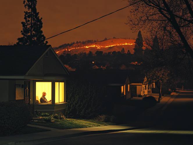 Elderly man visible in lit window as wildfire burns on hills behind dark California neighborhood during PSPS outage