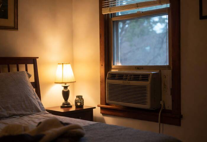 A cozy bedroom with a window air conditioner installed and a warm lamp beside the bed