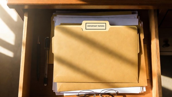 An open desk drawer with a plain manila folder, a pen and reading glasses
