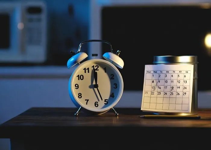 A twin-bell alarm clock and a small paper calendar on a kitchen table at dusk, a blank microwave display behind them