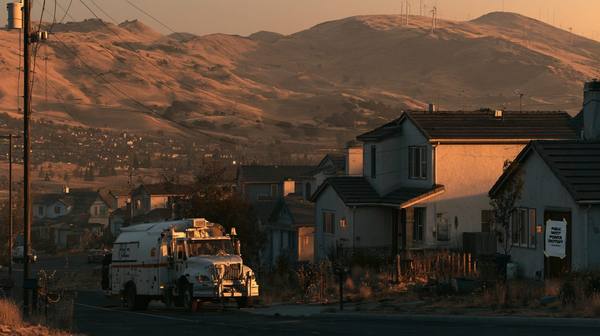 California neighborhood at dusk with a Public Safety Power Shutoff notice on a house and a PG&E utility truck parked on the street — golden dry hills in the background show the wildfire risk that triggers deliberate utility shutoffs