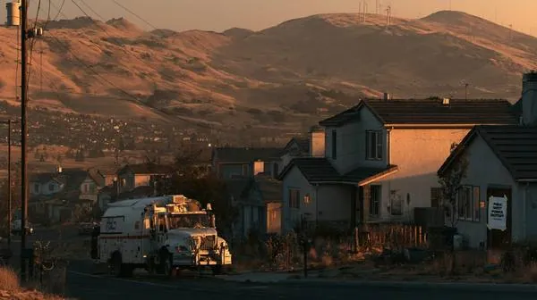 California neighborhood at dusk with a Public Safety Power Shutoff notice on a house and a PG&E utility truck parked on the street — golden dry hills in the background show the wildfire risk that triggers deliberate utility shutoffs