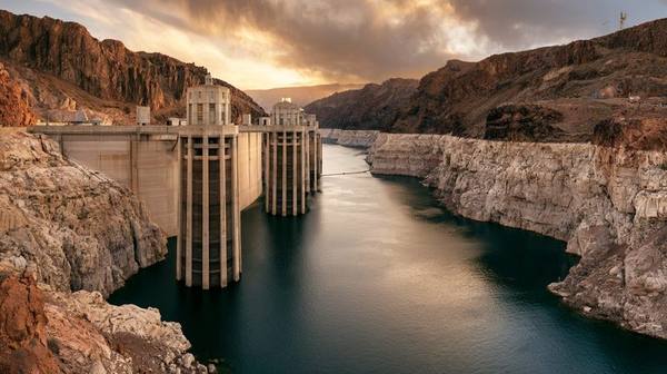 Hoover Dam intake towers rising from a dramatically low reservoir with exposed canyon walls showing the historic bathtub ring — prolonged drought has reduced hydroelectric generating capacity across the western United States