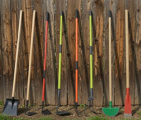 Lineup of long-handled garden tools — shovel, hoe, cultivator, and weeder — leaning against a wooden fence