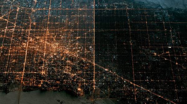 Satellite view looking straight down at a large city at night — left half illuminated with grid lights, right half completely dark during a rolling blackout showing how restoration priority leaves residential neighborhoods without power longest