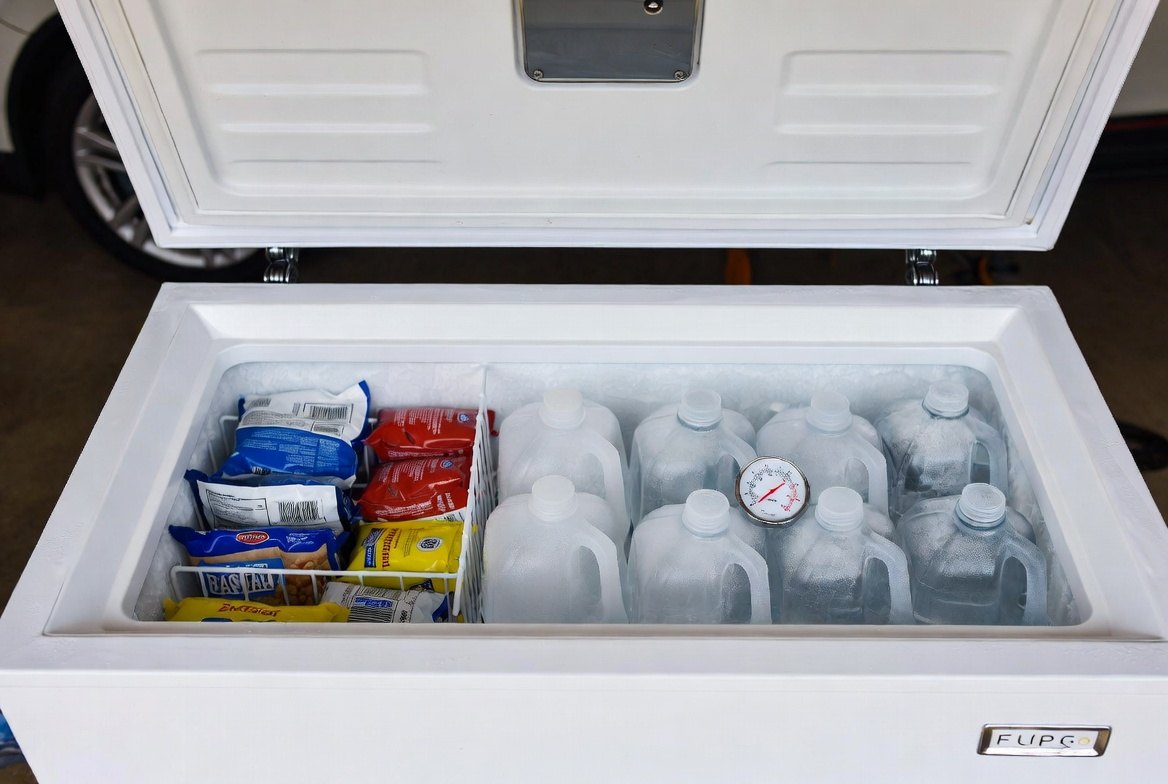 A chest freezer with its lid open, food packages in a basket on one side and rows of frozen one-gallon water jugs on the other, a dial thermometer resting on the jugs