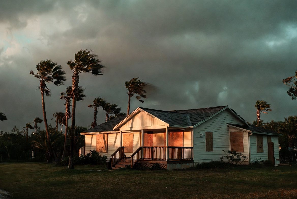 A single-story Florida house with plywood screwed over every window, palms bending in rising wind under a heavy storm sky at dusk