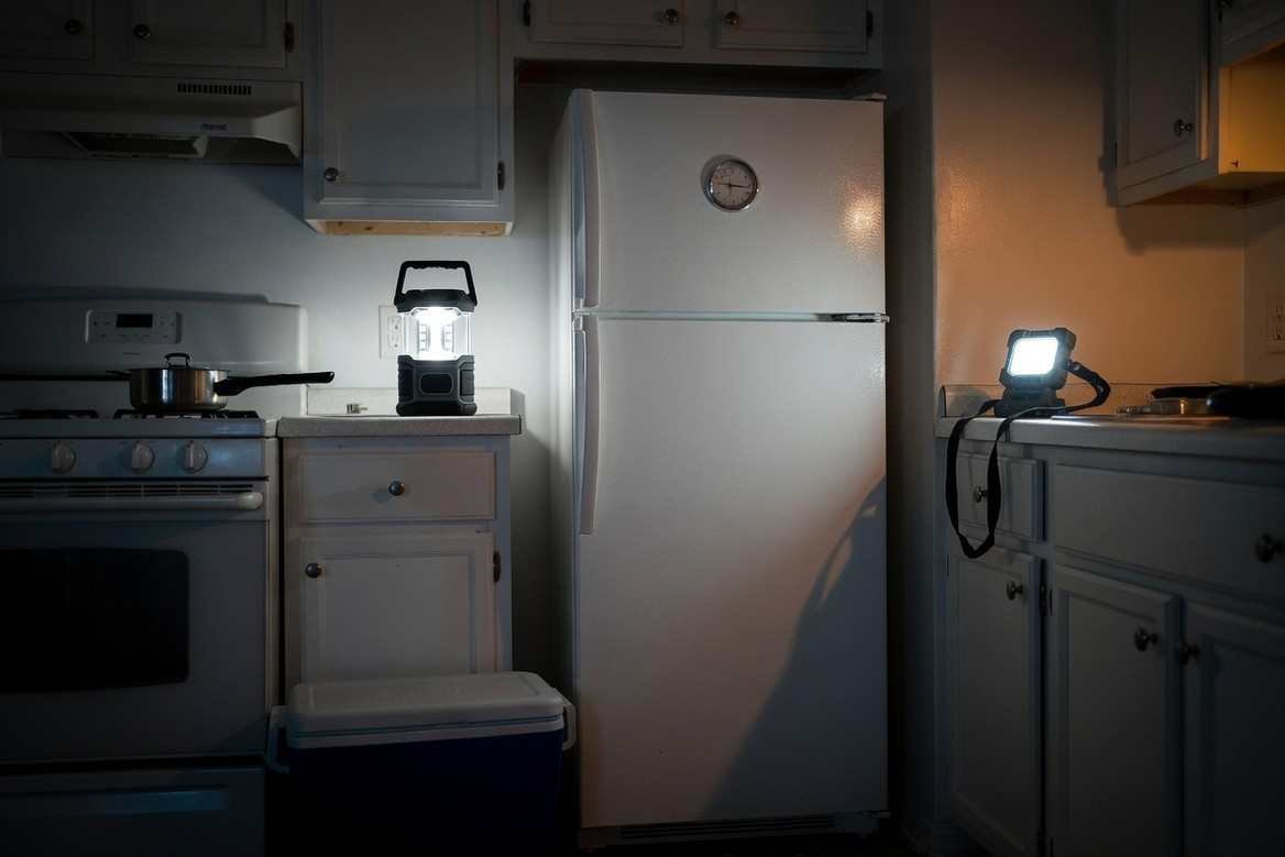 A closed refrigerator in a dark kitchen during a power cut, lit only by a battery lantern on the counter, with a cooler standing closed on the floor beside it