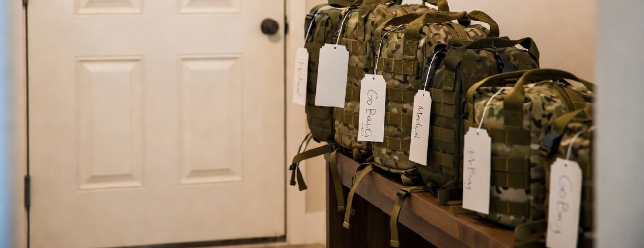 Emergency go-bags of different sizes with name tags sitting on a bench beside a front door, ready to grab and go