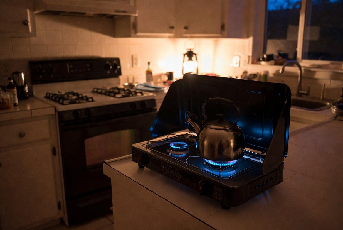 A small two-burner camp stove lit on a kitchen counter with a kettle on it during a power cut, the electric range beside it dark and unused, a lantern glowing behind