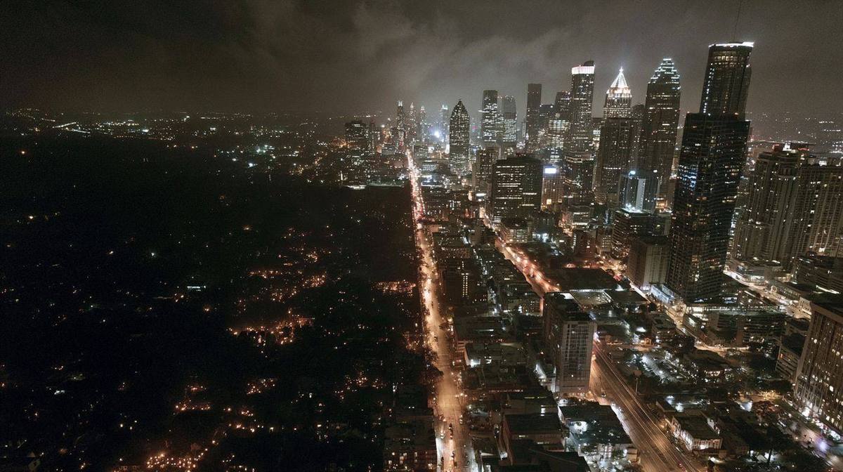 Aerial view of a major American city at night with residential neighborhoods completely dark and downtown still lit showing the restoration priority queue after a major power outage