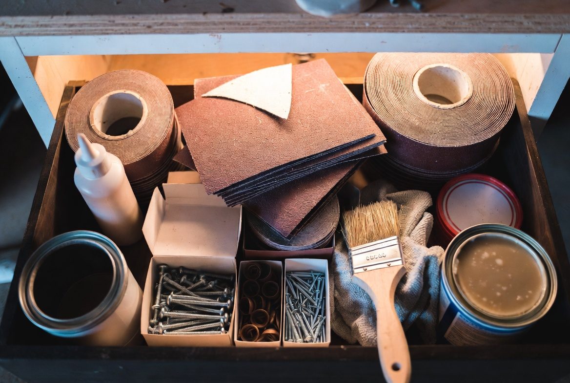 An open workshop drawer seen from above holding rolls and sheets of sandpaper, a glue bottle, boxes of screws and nails, a paintbrush on a rag and two open tins of finish
