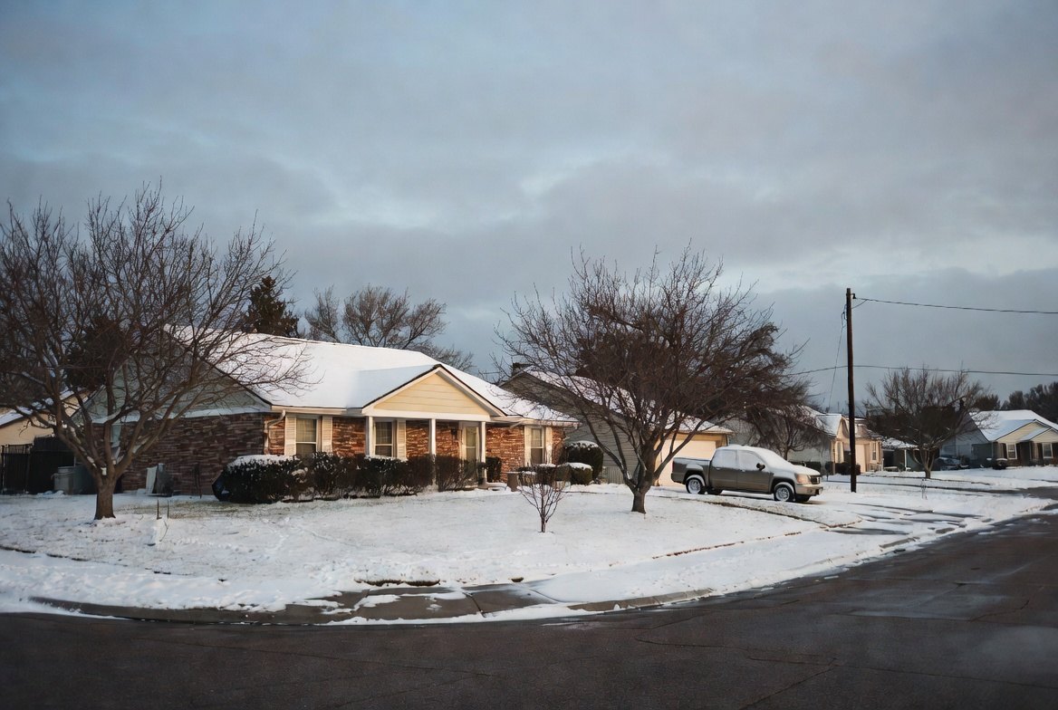 A snow-covered street of single-story Texas brick houses under a heavy grey sky, bare trees and overhead power lines, one pickup truck in a driveway and no one outside