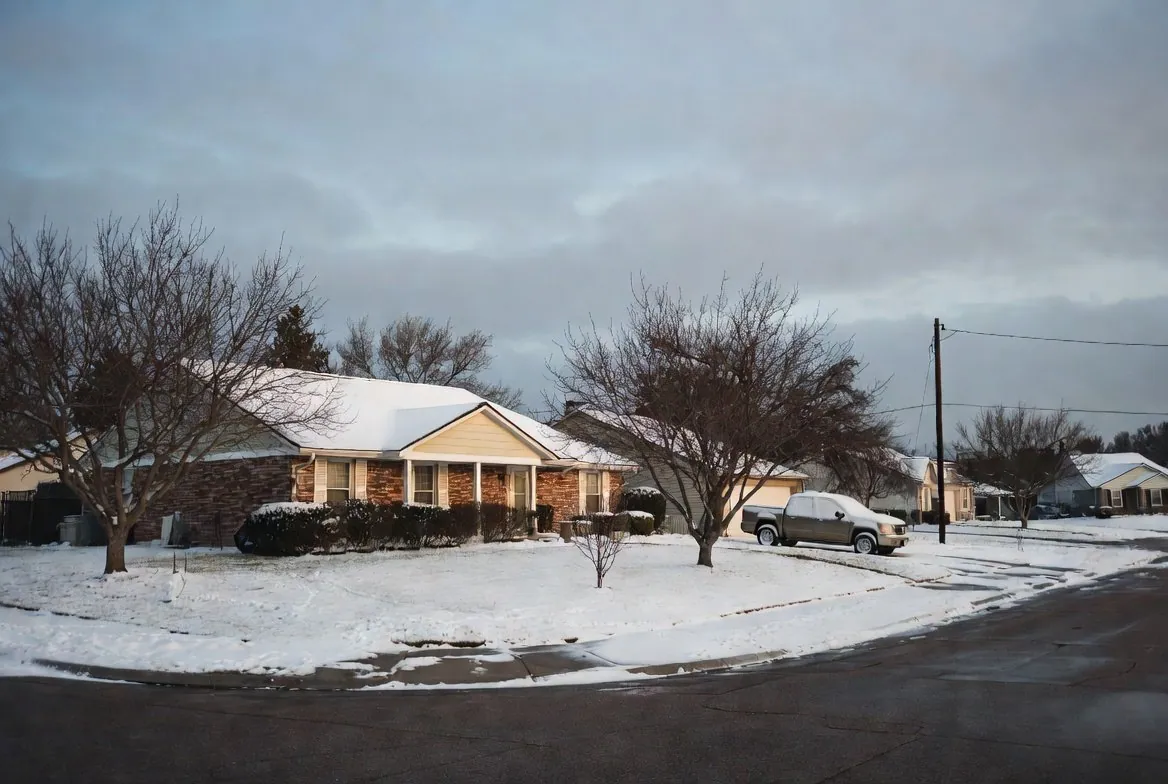 A snow-covered street of single-story Texas brick houses under a heavy grey sky, bare trees and overhead power lines, one pickup truck in a driveway and no one outside