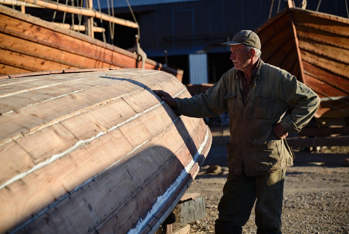 An older boatwright in a boatyard resting one hand flat on the planking of an upturned wooden hull in low golden light