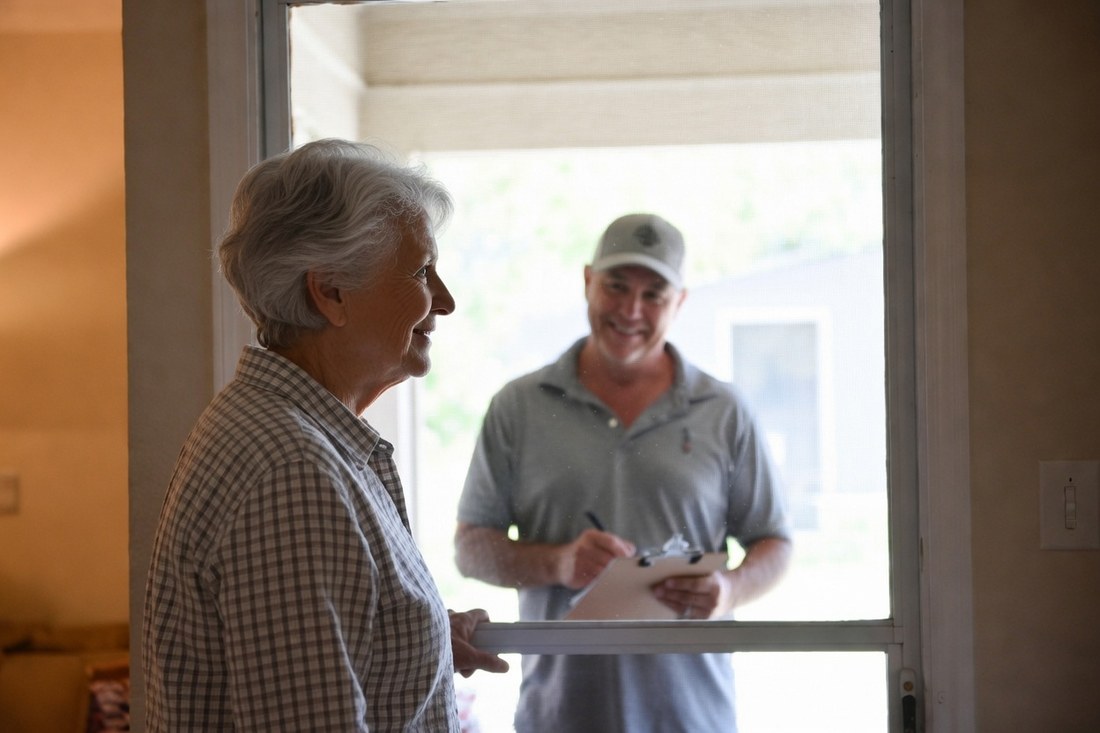 A silver-haired woman inside her home speaking politely through a closed screen door to a smiling man in a gray polo and ball cap holding a clipboard on the porch