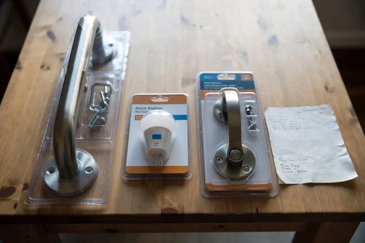 Four things laid out on a wooden table: a boxed stainless steel grab bar, a plug-in LED night light, a lever door handle, and a handwritten list on a sheet of notepaper