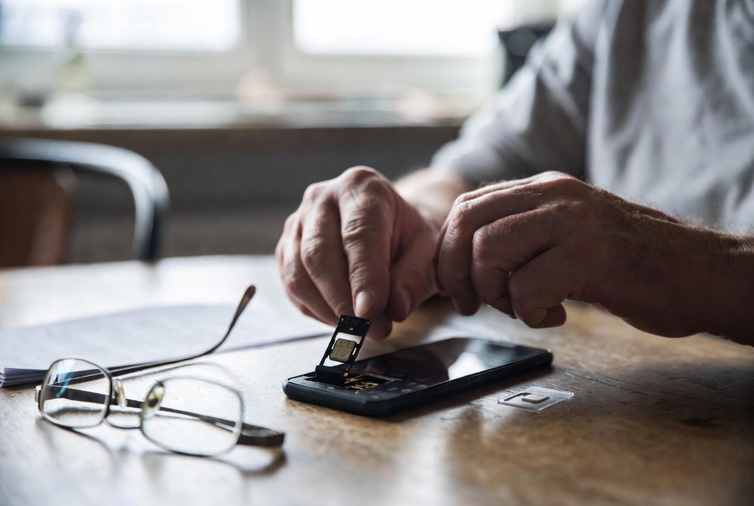 An older personβs hands at a kitchen table easing the SIM tray out of a plain black phone, with a new SIM card in its carrier on the table and reading glasses beside it
