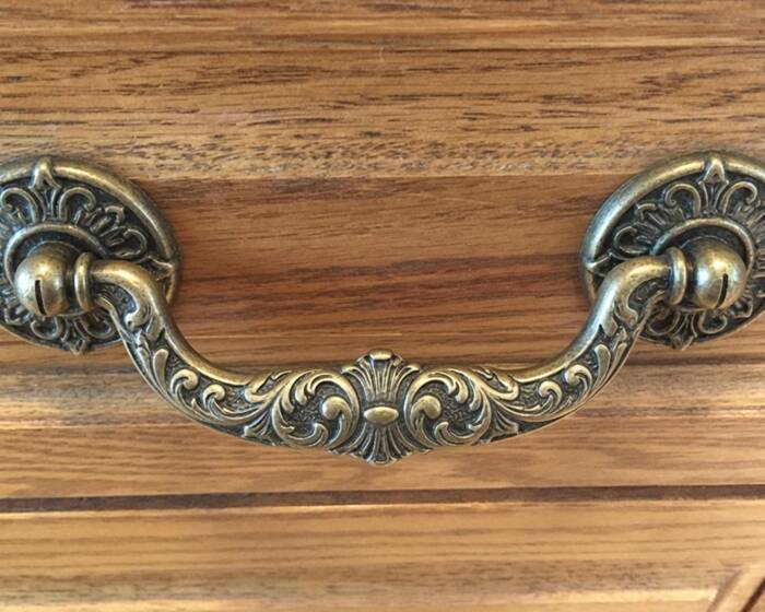 An ornate antique brass drop bail pull on a wooden drawer, with room to hook fingers underneath the swinging handle