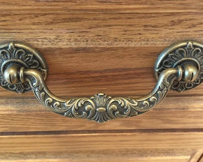 An ornate antique brass drop bail pull on a wooden drawer, with room to hook fingers underneath the swinging handle