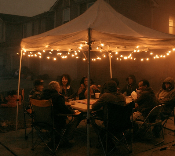 Neighbors gathered under a white pop-up canopy tent lit with string lights at night, sitting around a folding table in lawn chairs