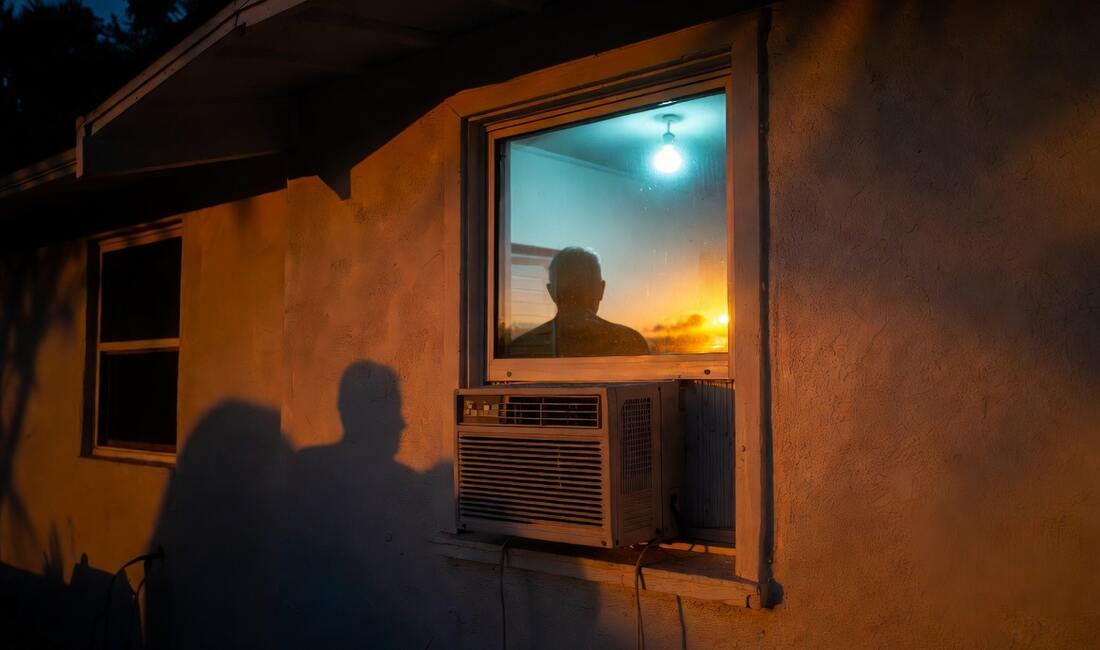 A window air conditioner in the lit window of a modest home at dusk, a man's silhouette inside
