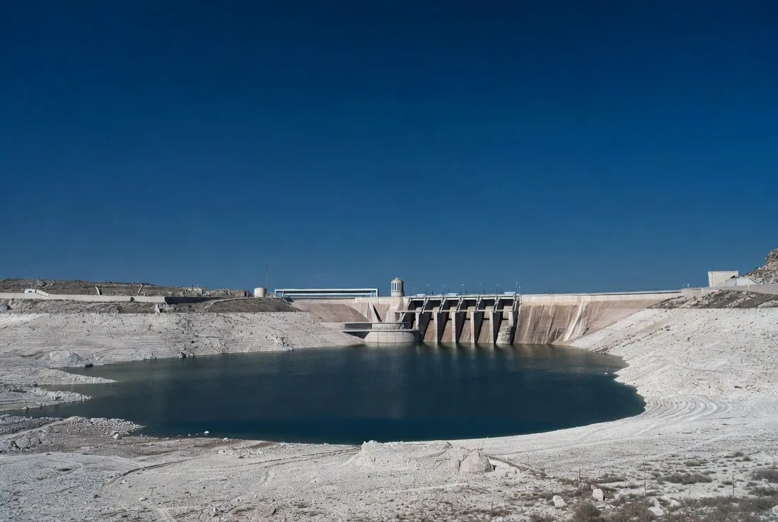 A large concrete hydroelectric dam with the reservoir far below its normal level, a wide pale bathtub ring of bleached rock exposed around the shoreline