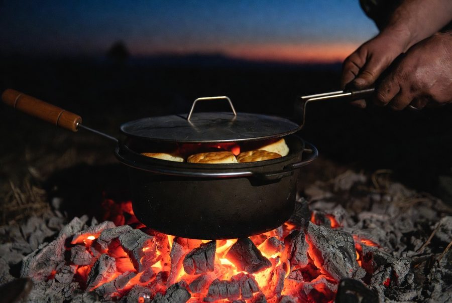 The lid of a camp Dutch oven lifted away with a lid lifter tool at dusk, revealing golden-brown biscuits baking inside over glowing coals