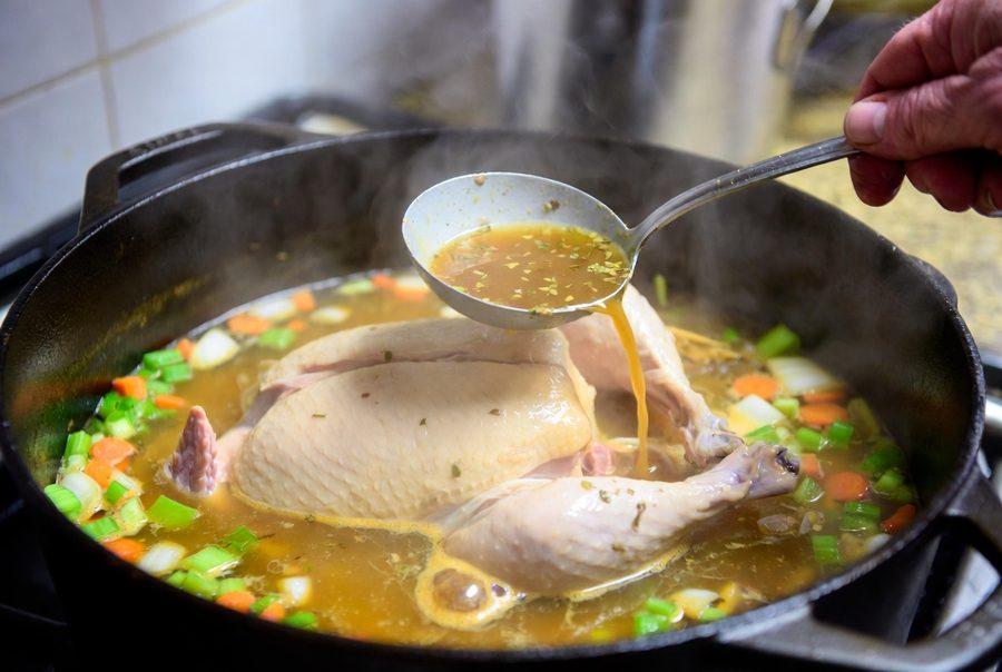 A whole chicken stewing in golden broth with carrots, celery, and onions in a cast iron Dutch oven, a ladle basting broth over the bird