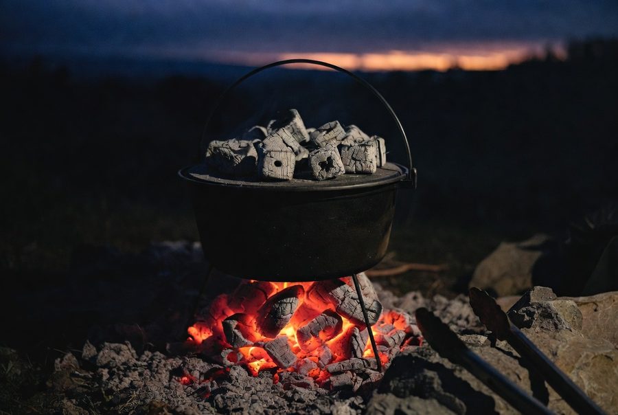A three-legged camp Dutch oven at dusk standing over glowing coals with charcoal briquettes arranged on its flanged lid, tongs resting nearby