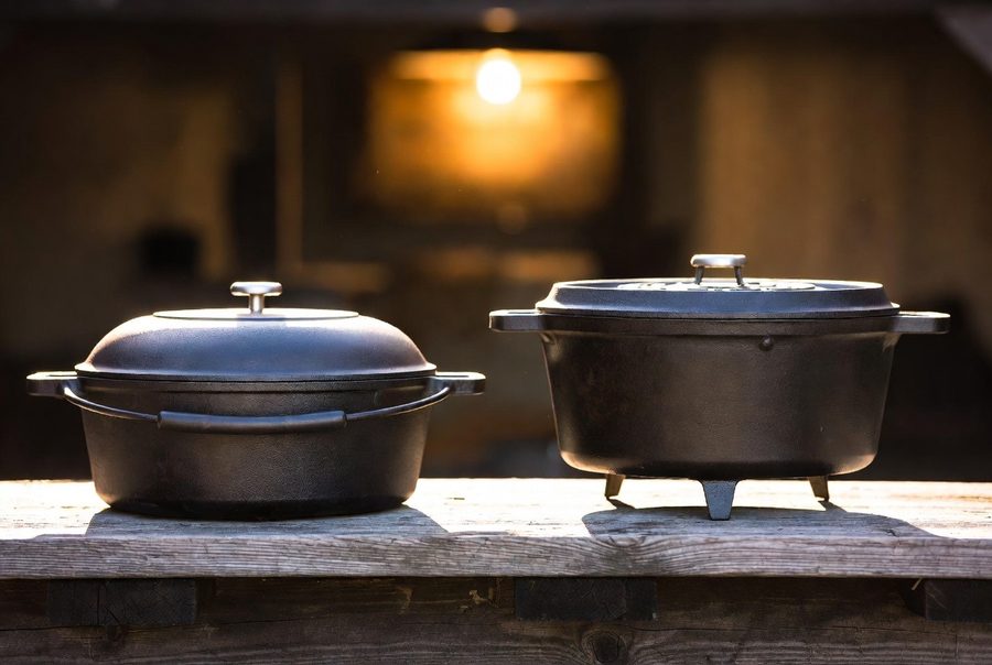 The two branches side by side on weathered wood: a flat-bottom kitchen Dutch oven and a three-legged camp Dutch oven built to stand over coals