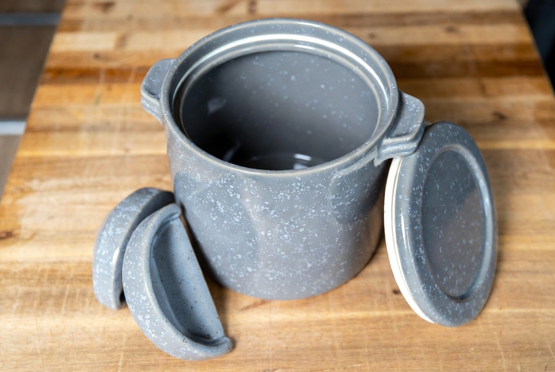 A grey stoneware fermenting crock on a wooden board with its lid off, showing the water channel around the rim, and two heavy crescent-shaped stone weights leaning against its base