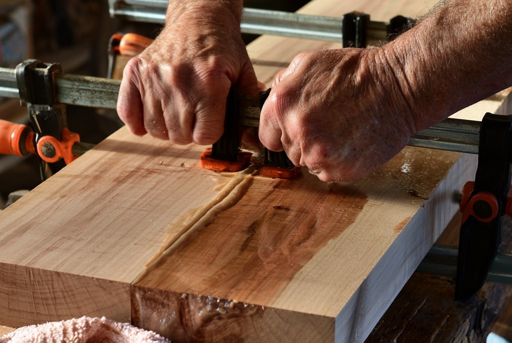 Two boards clamped edge to edge on a workbench with bar clamps, a bead of glue squeezed out along the joint, older hands tightening the clamps and a damp cloth beside them