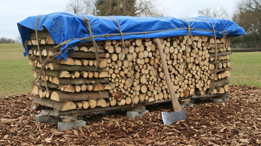 A long row of split firewood stacked on pallets raised on concrete blocks in an open field, with a tarp tied over the top of the stack only and both sides left fully open to the air