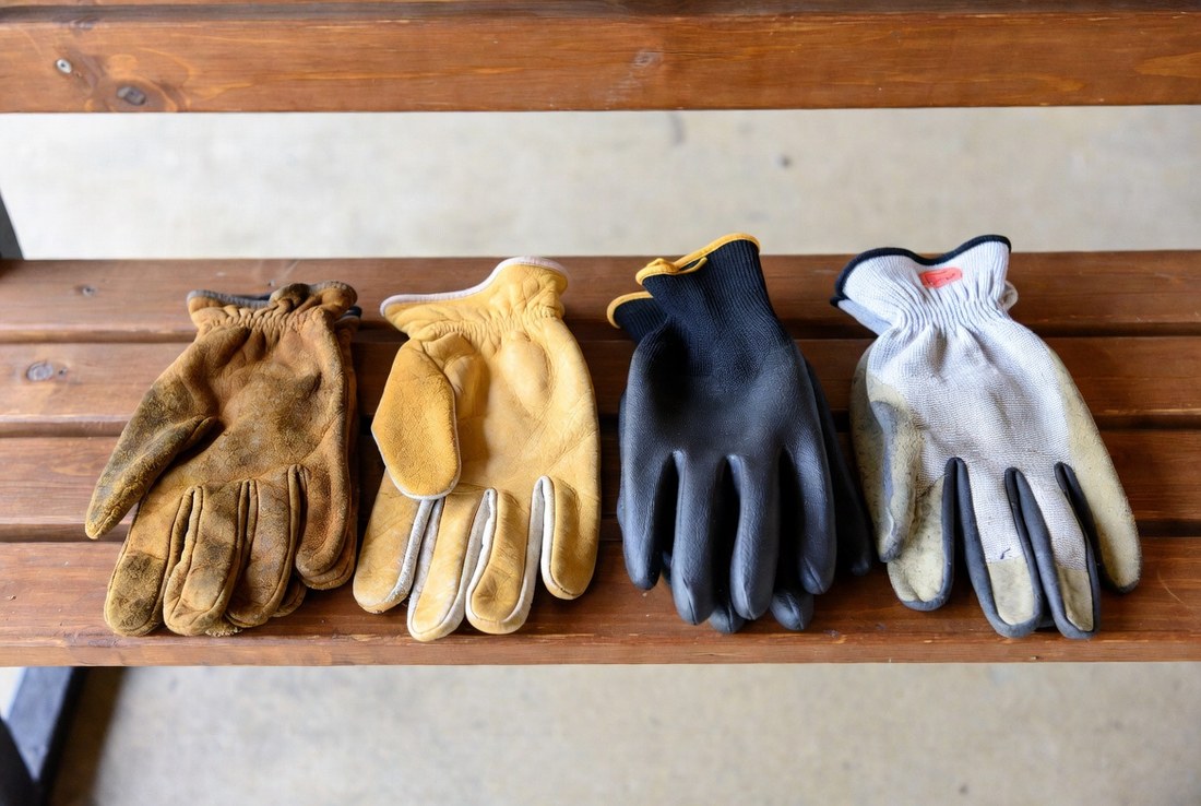 Four pairs of work gloves laid side by side on a wooden bench — worn heavy leather, pale goatskin, black nitrile-coated knit and a lighter coated knit pair
