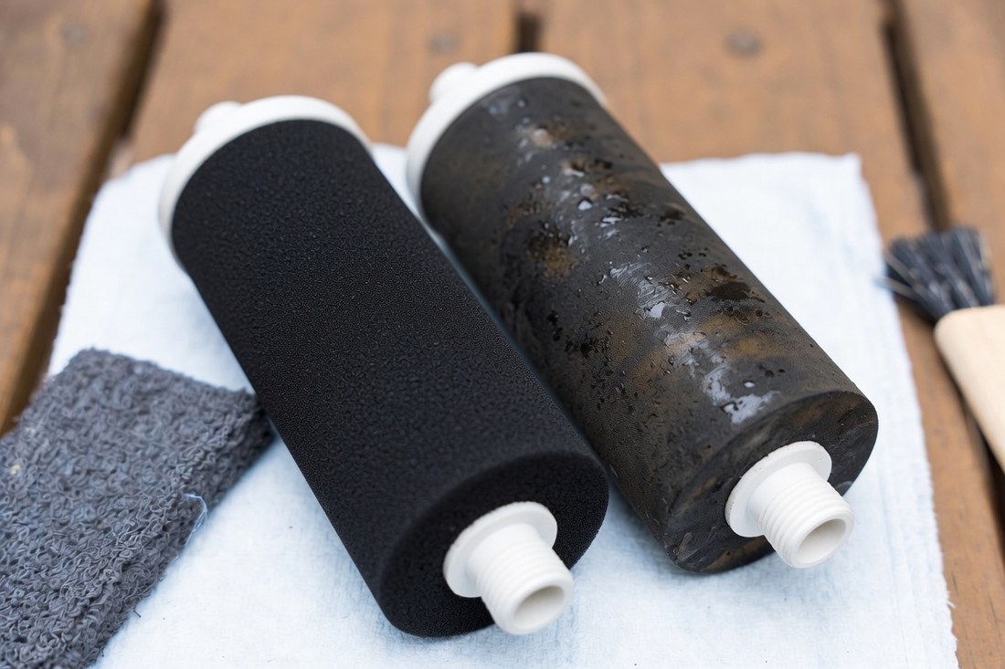 Two black cylindrical filter elements lying on a white cloth, one clean and matte, the other darkened and wet with fouling, a scrubbing pad and brush beside them