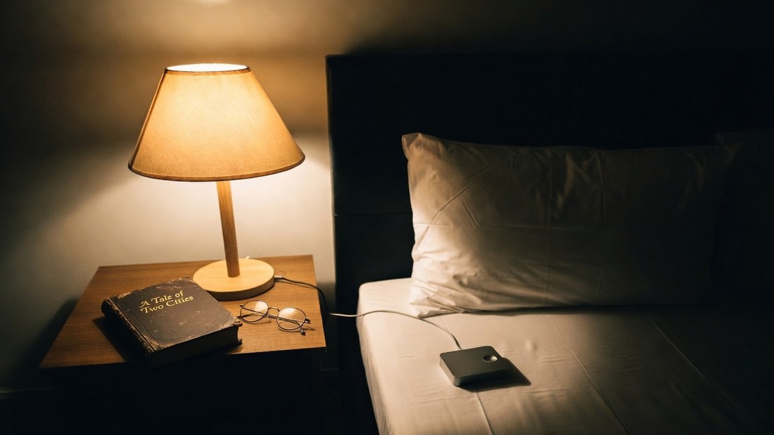 A bedside table at night with a lit lamp, reading glasses and a worn hardback copy of A Tale of Two Cities, and a small bed-shaker unit lying on the mattress with its cable running back to the table