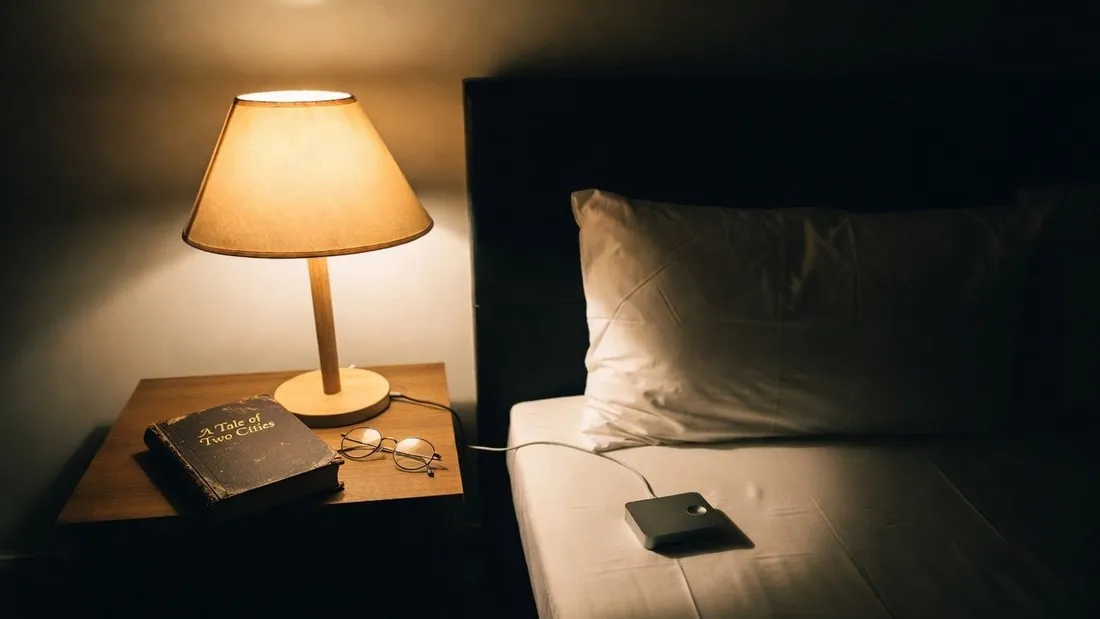 A bedside table at night with a lit lamp, reading glasses and a worn hardback copy of A Tale of Two Cities, and a small bed-shaker unit lying on the mattress with its cable running back to the table