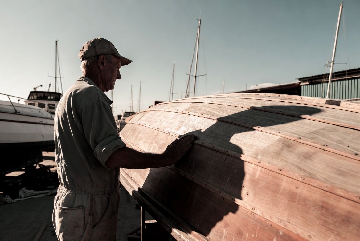 A man in work coveralls standing beside a wooden hull in a marina, one hand laid flat against the planking
