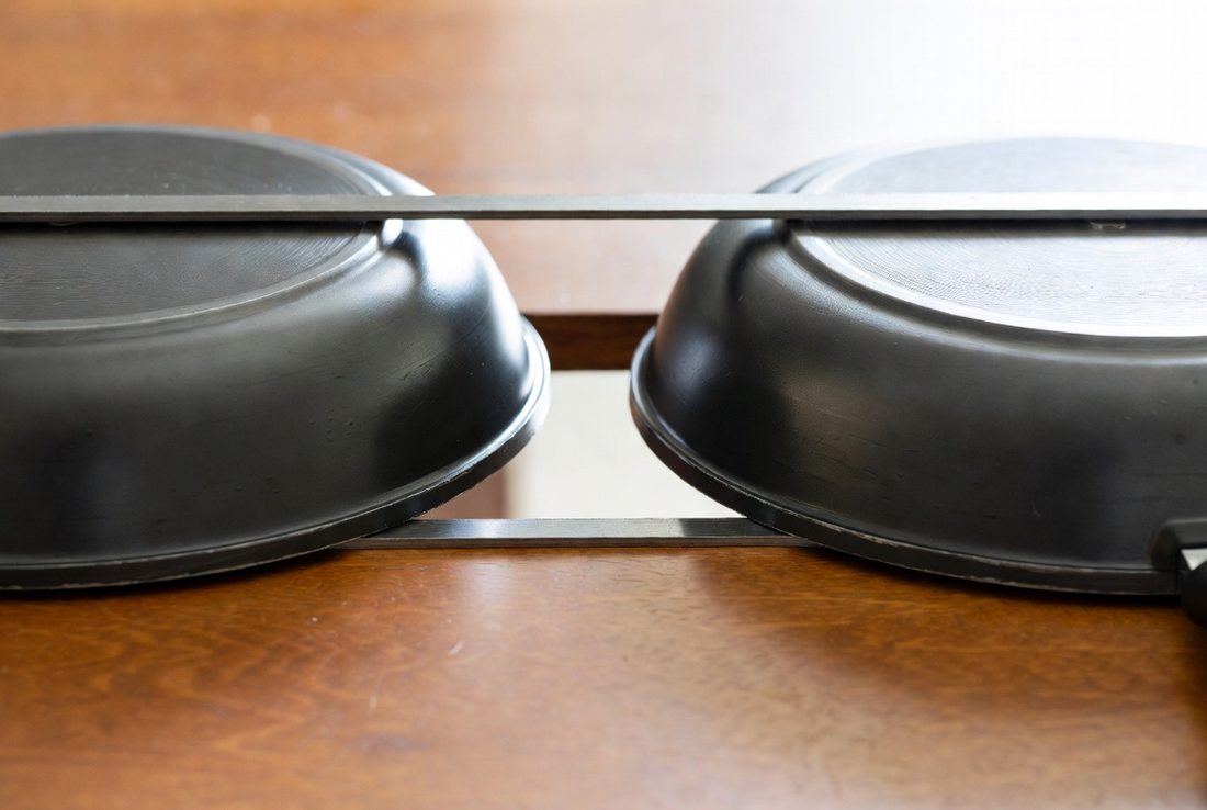 Two skillets turned upside down on a wooden table with their bases uppermost and a steel straightedge laid across them, photographed at table level so the line of contact is visible