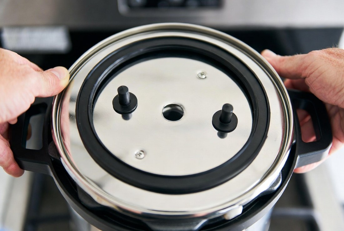 The underside of an electric pressure cooker lid held in two hands, the silicone sealing ring seated in its groove and two valve posts standing up from the plate