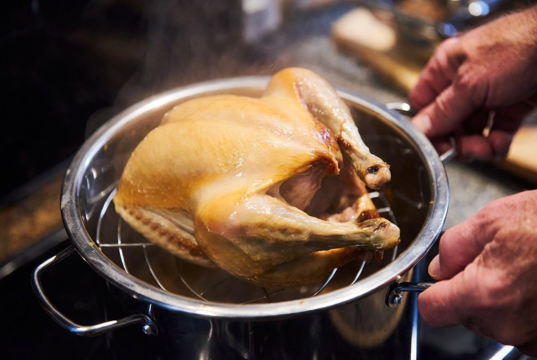 A whole cooked chicken being lifted out of a pressure cooker on a wire trivet, steam rising, hands on the trivet handles