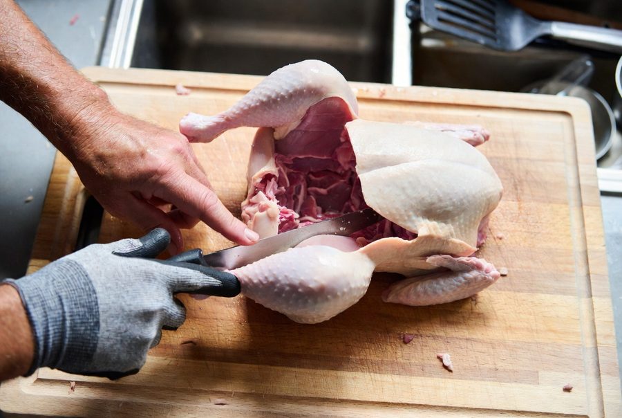 Overhead view of hands breaking down a whole raw chicken with a boning knife, a gray cut-resistant glove on the holding hand