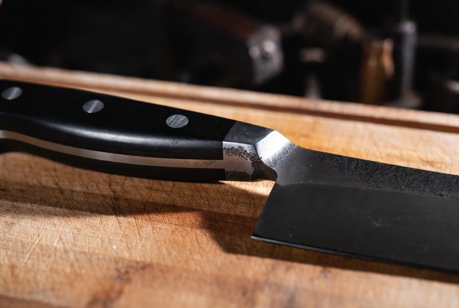 Macro close-up of a forged chef's knife showing the thick bolster and the full tang running through the riveted handle