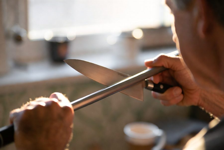 An older man drawing a chef's knife along a honing steel at a shallow angle in warm morning window light