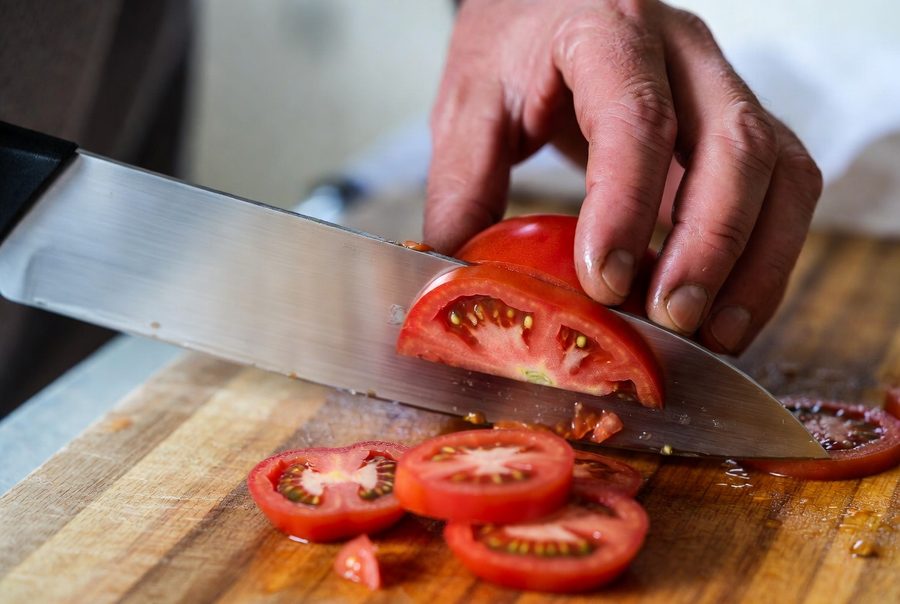 A sharp chef's knife slicing cleanly through a ripe tomato, thin slices already cut, the blade biting the skin on the first pass