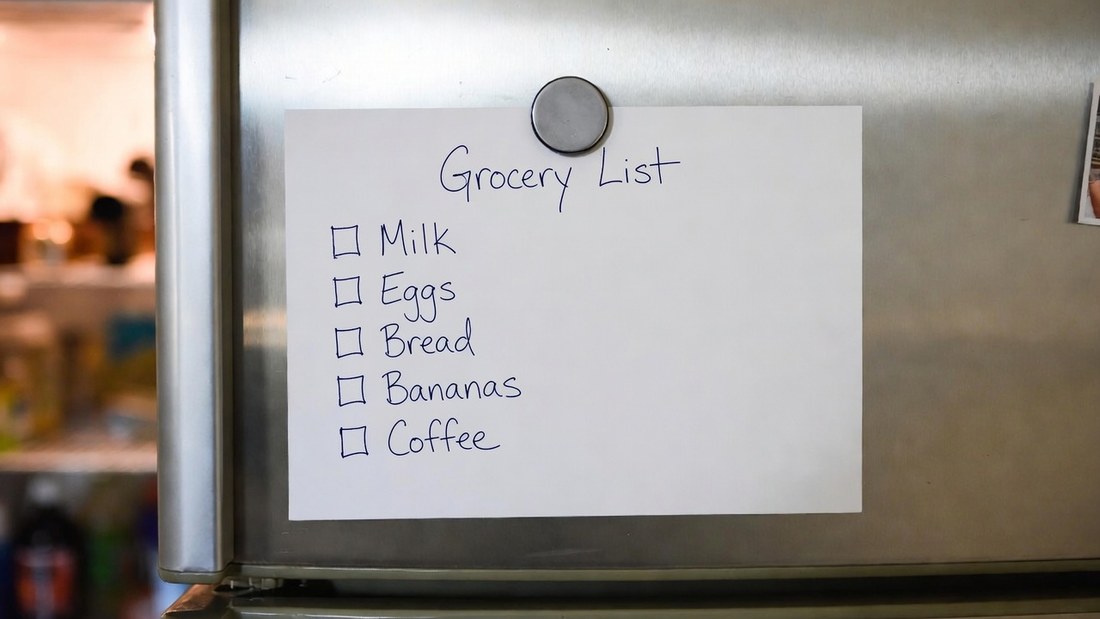 A handwritten grocery list held to a stainless refrigerator door by a round magnet
