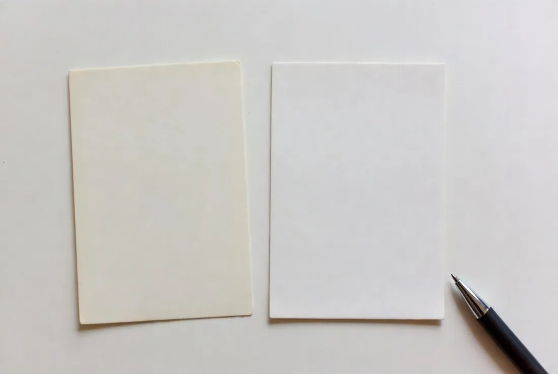 Two blank index cards lying side by side on a plain surface, one yellowed and soft at the corners and one crisp and new, a pen beside them