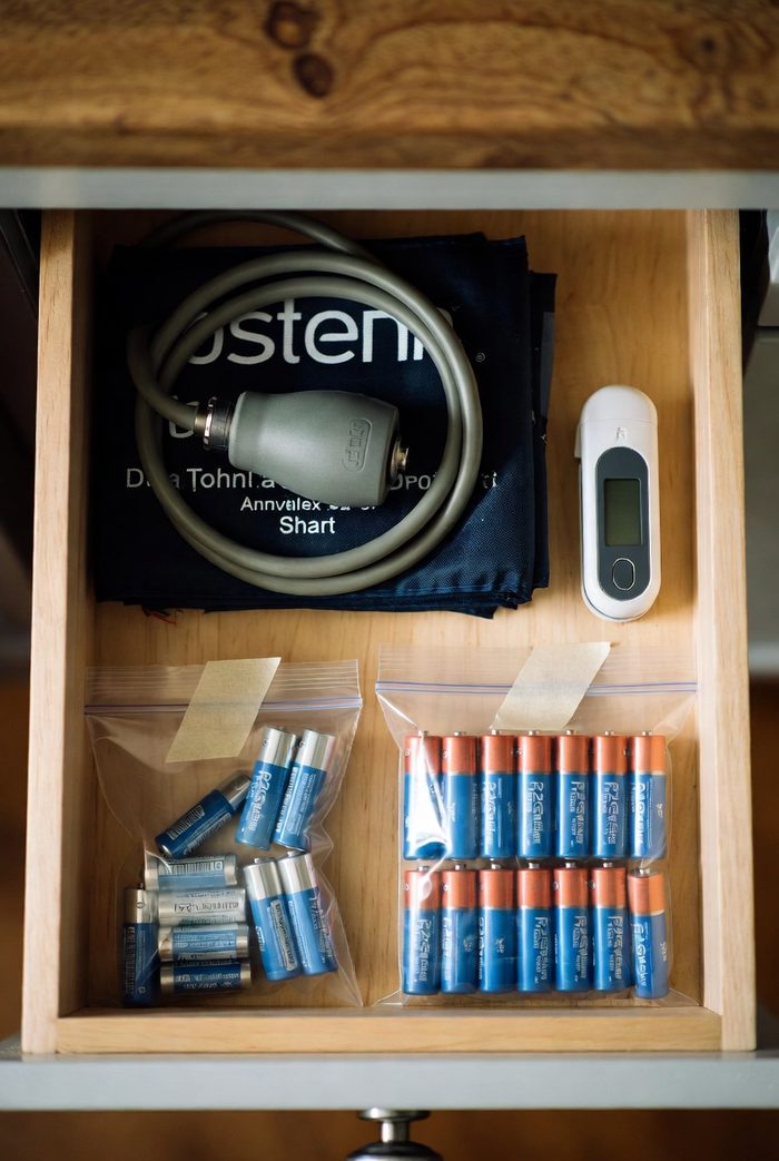 Overhead view of an organized drawer: blood pressure cuff, thermometer, and two zip bags of spare batteries taped with masking tape
