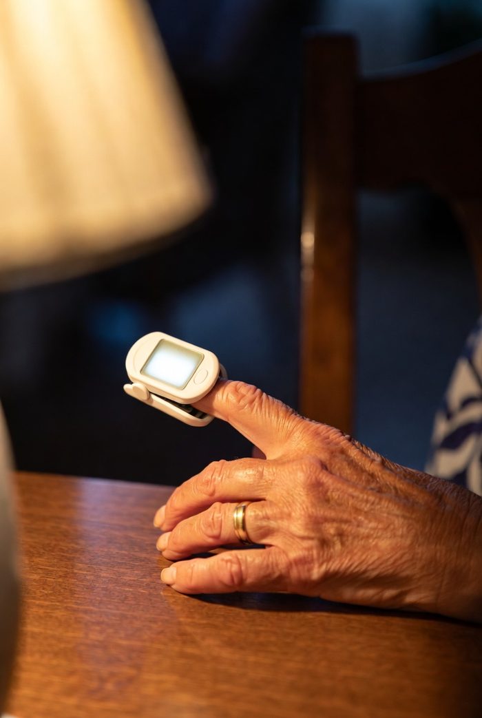 Older woman's hand on a wooden table with a white fingertip pulse oximeter clipped on her finger in warm lamp light